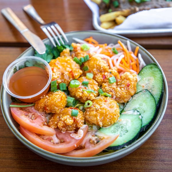A Vietnamese bowl with crispy shrimp, sliced cucumbers, tomatoes, pickled carrots, and a side of sauce.