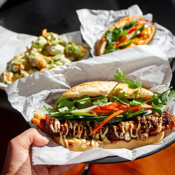 A hand holds a Banh Mi Boys banh mi sandwich with crispy chicken, cilantro, and vegetables.
