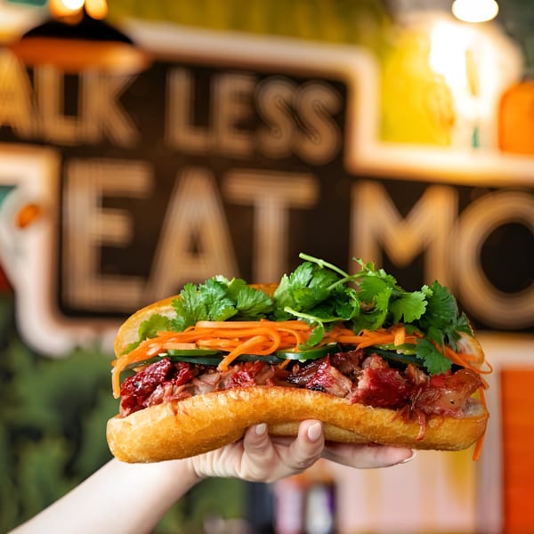 A hand holds a Banh Mi Boys banh mi sandwich filled with meat, cilantro, and pickled vegetables.