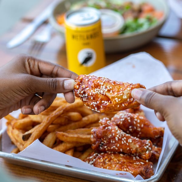 Hands picking up a sesame-crusted chicken wing from a tray of wings and fries, with a drink in the background.