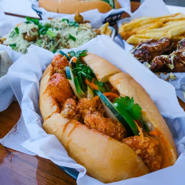 A Banh Mi Boys banh mi sandwich with crispy chicken, vegetables, and sauce, alongside fries and wings.