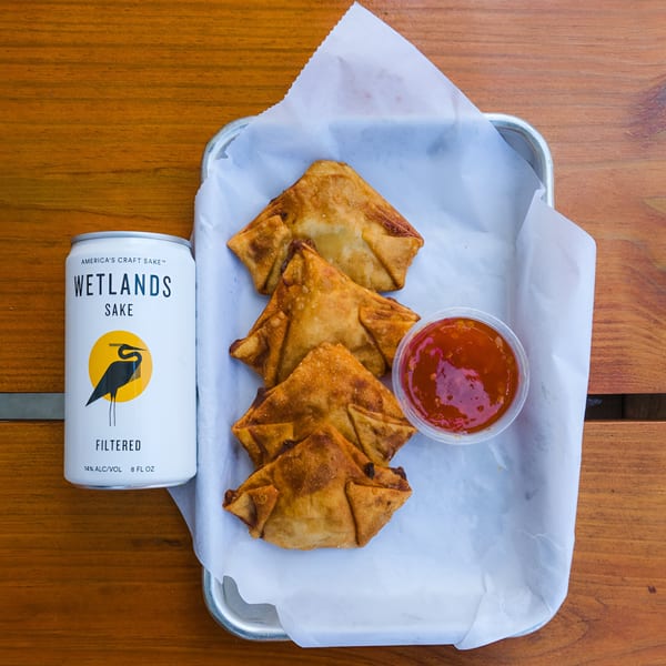 Crispy fried wontons with sweet chili sauce and a can of Wetlands Sake on a wooden table.