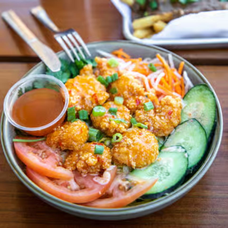 A bowl of Banh Mi Boys' crispy shrimp with sliced cucumbers, tomatoes, and a side of sauce.